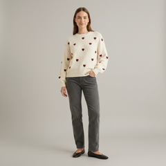 Woman wearing cream knit pullover with scattered heart pattern, drop shoulder sleeves, relaxed fit, neutral backdrop