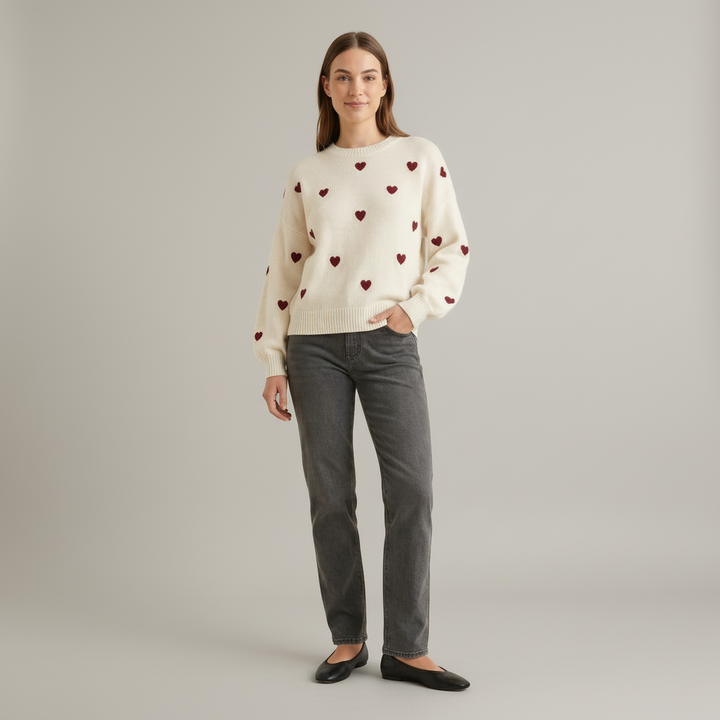 Woman wearing cream knit pullover with scattered heart pattern, drop shoulder sleeves, relaxed fit, neutral backdrop